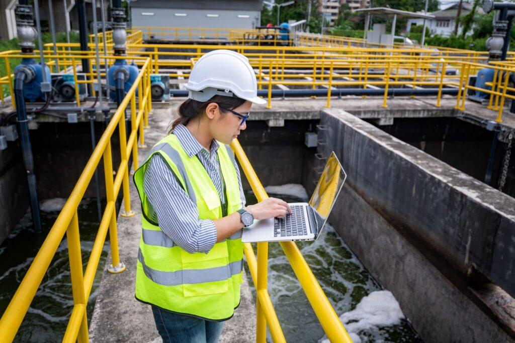 Eine Ingenieurin mit Schutzhelm und Warnweste arbeitet an einem Laptop in einer Wasseraufbereitungsanlage. Sie überwacht den Betrieb der Anlage, während im Hintergrund Rohre, Geländer und industrielle Ausrüstungen zu sehen sind. Die Szene zeigt den Einsatz moderner Technik zur Sicherstellung einer effizienten Wasseraufbereitung.