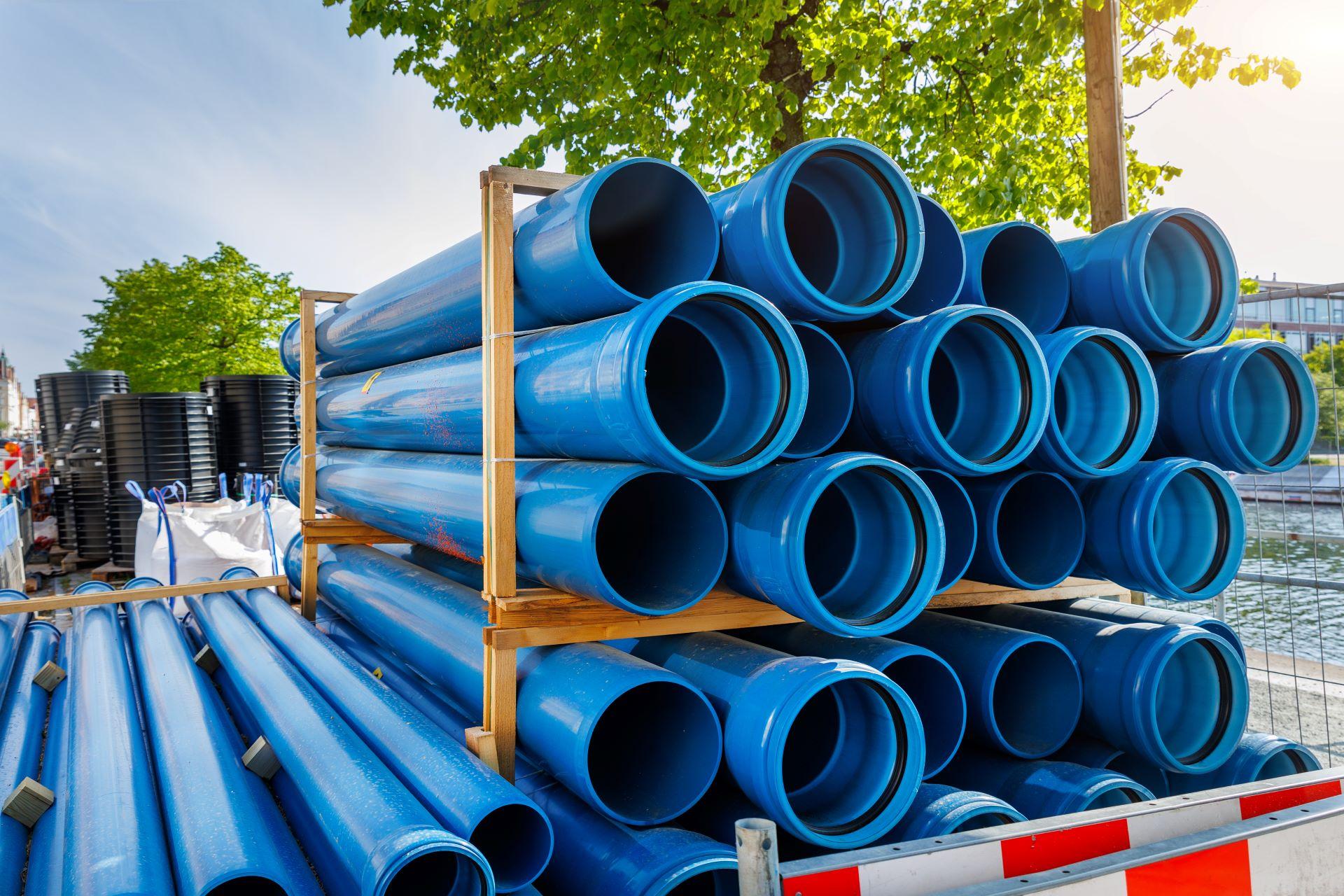 Ein Stapel blauer GFK-Rohre auf einer Baustelle, die bereit zur Installation sind. Die Rohre sind ordentlich auf einem Holzgestell gestapelt, während im Hintergrund weitere Baumaterialien und Bäume bei sonnigem Wetter zu sehen sind.