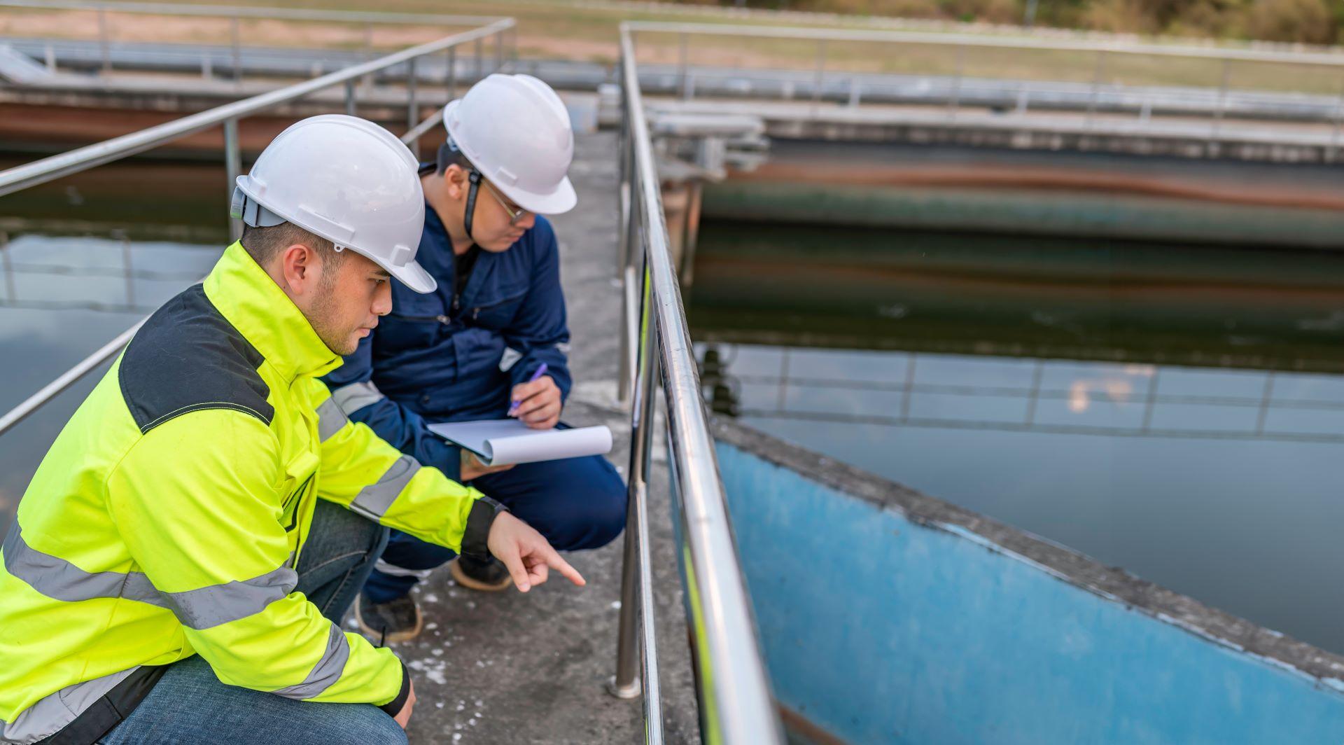 Zwei Ingenieure mit Schutzhelmen und Sicherheitskleidung inspizieren eine Wasseraufbereitungsanlage. Sie konzentrieren sich auf die Überprüfung der Infrastruktur und die Sicherstellung einer effizienten und umweltfreundlichen Wasserversorgung.